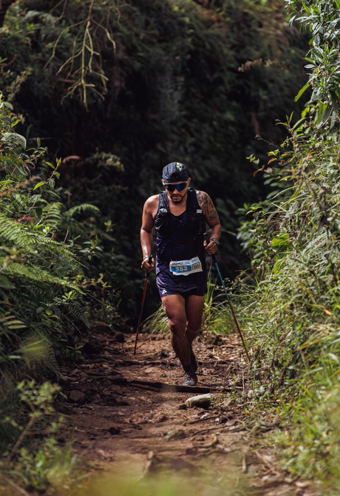 Coureur de trail muni de bâtons sur un sentier étroit, au milieu d'une végétation dense qui ferme l'horizon.